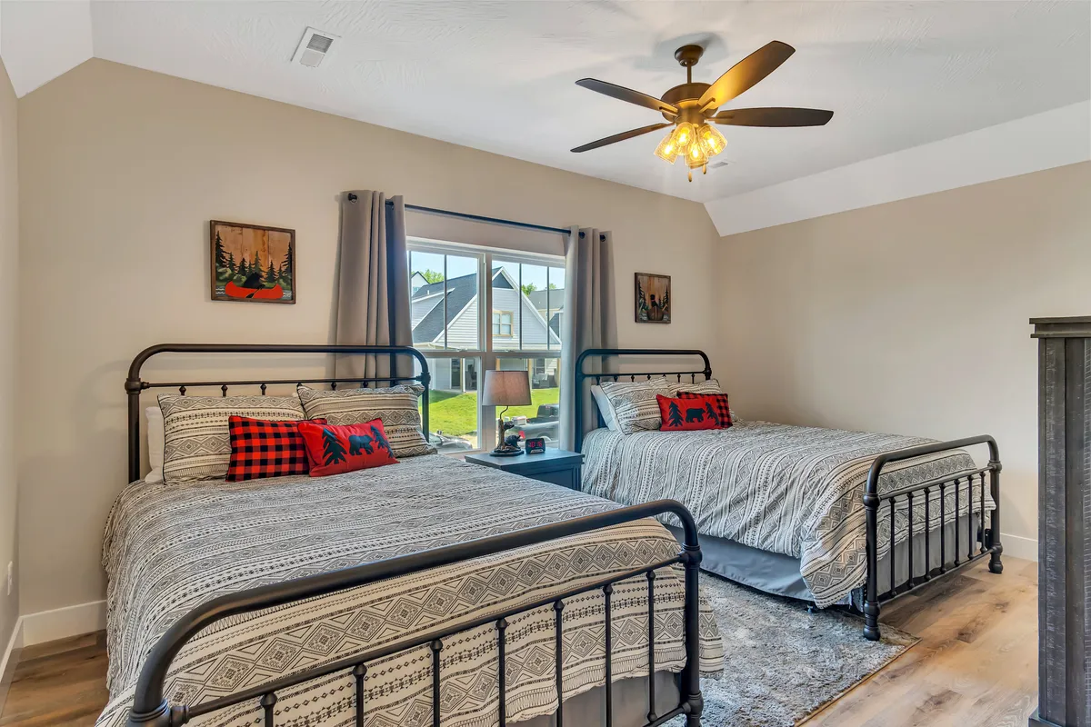 A spacious bedroom with two neatly made beds adorned with decorative pillows. Natural light pours in from a large window, and a ceiling fan with a light fixture adds to the room’s comfortable, welcoming feel. The setup is perfect for friends or family sharing a room.