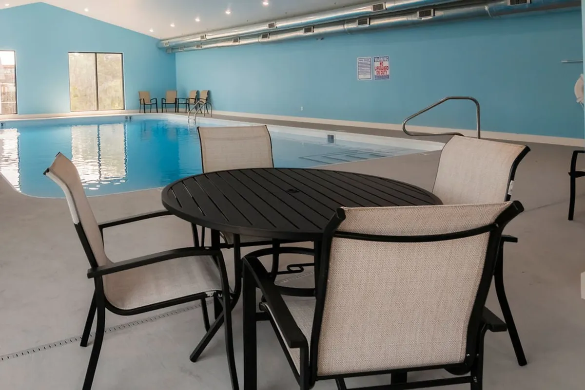 A bright, airy indoor swimming pool area with natural light streaming in through large windows. The space includes a round table with chairs for relaxing and socializing, reinforcing a sense of community and year-round enjoyment.
