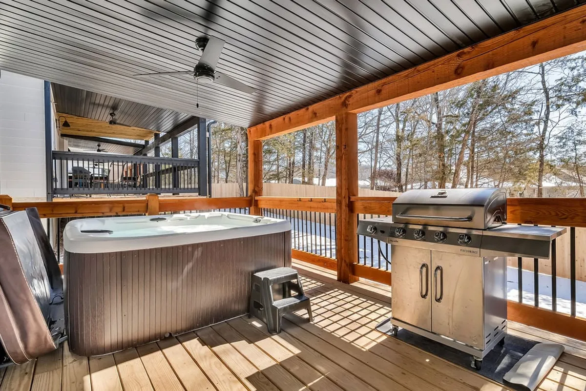 A well-appointed wooden deck featuring a private hot tub, a modern grill, and an inviting deck area surrounded by lush trees. The space is perfect for morning coffee, a relaxing soak, or an evening barbecue.