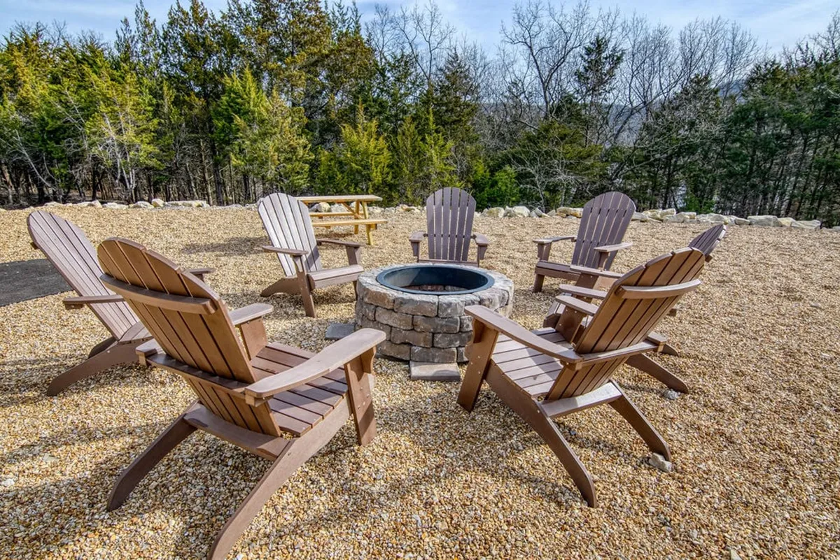 A cozy outdoor fire pit encircled by Adirondack chairs and flanked by tall trees, suggesting evenings spent with friends and family, storytelling, and stargazing under the night sky.
