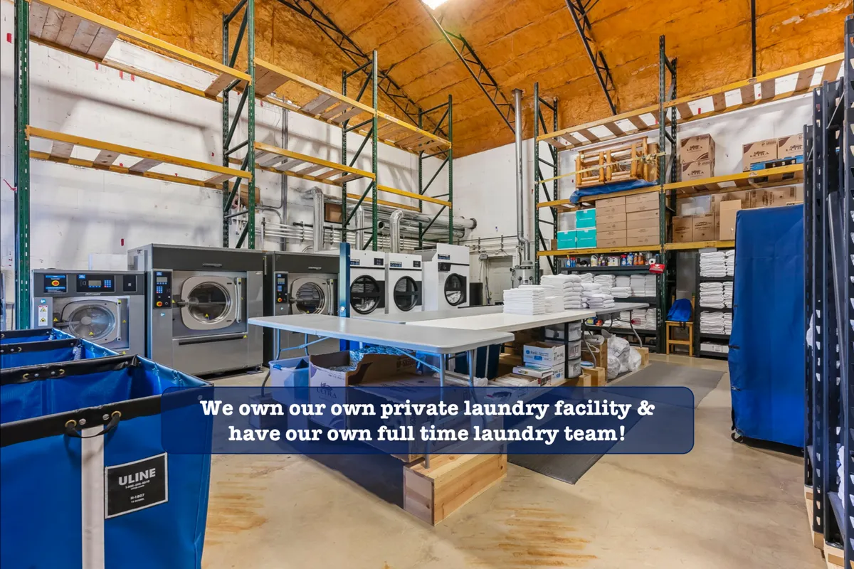A behind-the-scenes look at an industrial laundry setup, featuring commercial washing machines, a folding table, and storage shelves with supplies. The image conveys the scale and professionalism of the cleaning processes.