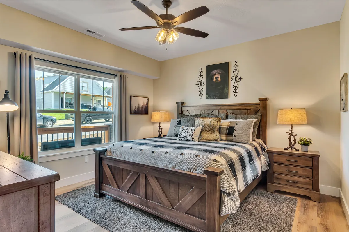 A king-sized bed with inviting plaid bedding is flanked by wooden nightstands, while a large window lets in abundant natural light. The room is decorated with a playful dog artwork on the wall, adding a touch of personality. The setting is peaceful, comfortable, and ideal for restful nights.