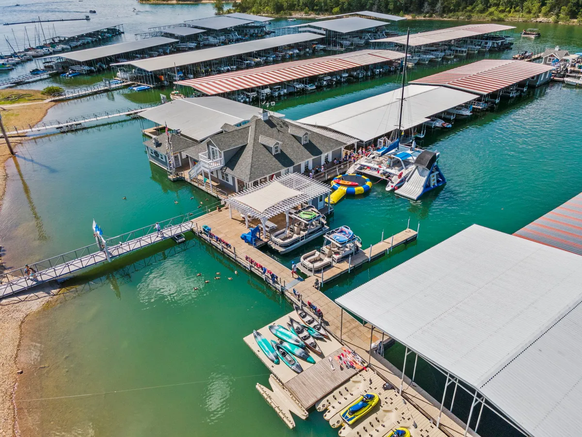 A lively waterfront dock scene with a boathouse, watercraft, and people enjoying boating, kayaking, and swimming. The background features calm water and the social energy of friends gathering for water-based fun, with sunset hues enhancing the atmosphere.