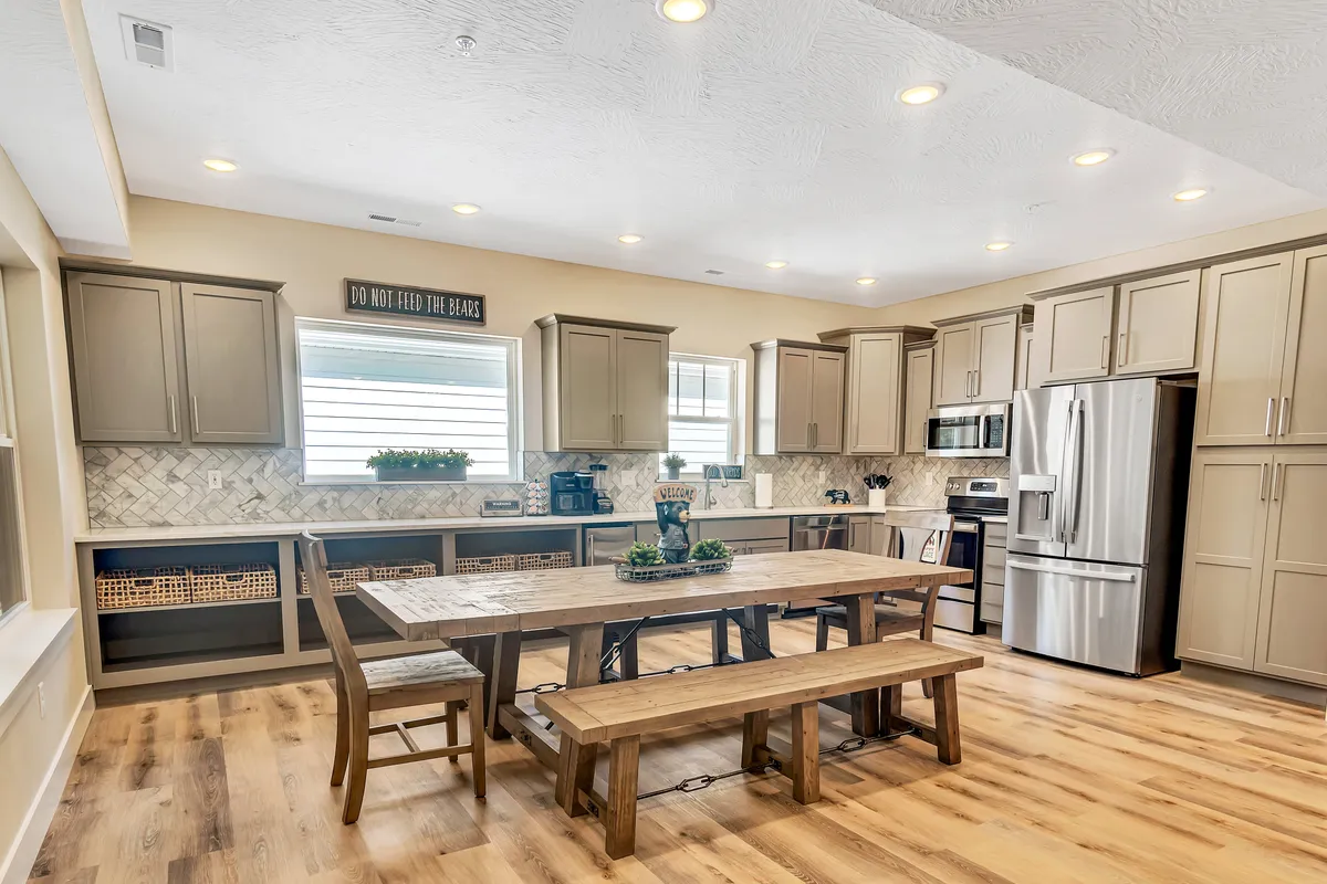 This image reveals a spacious dining area with a large wooden table, modern kitchen appliances in the background, and sunlight streaming through an open window with greenery outside. The scene radiates a sense of togetherness, ideal for family meals, celebrations, or casual breakfasts.