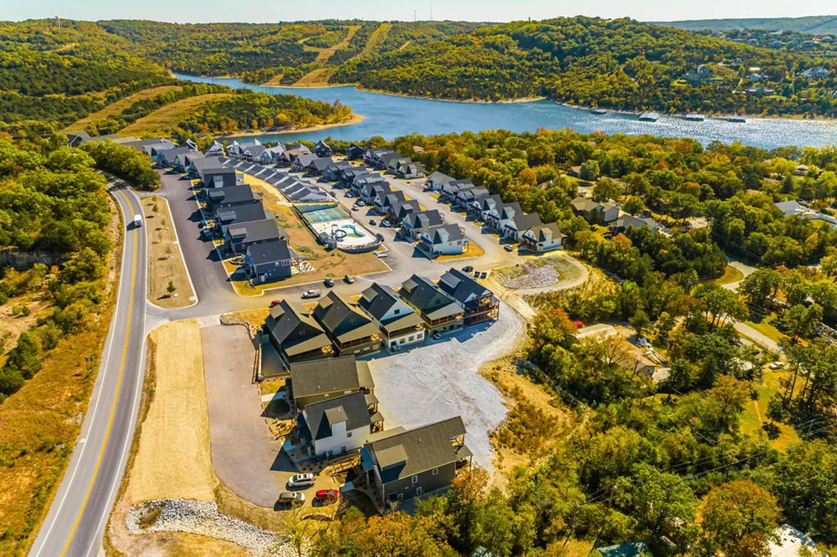 A peaceful view of Table Rock Lake with a cluster of houses nearby, surrounded by lush greenery and a winding road. The lake is calm, perfect for boating, swimming, or relaxing by the water. The image captures the expansive shoreline and the inviting natural beauty of the area.